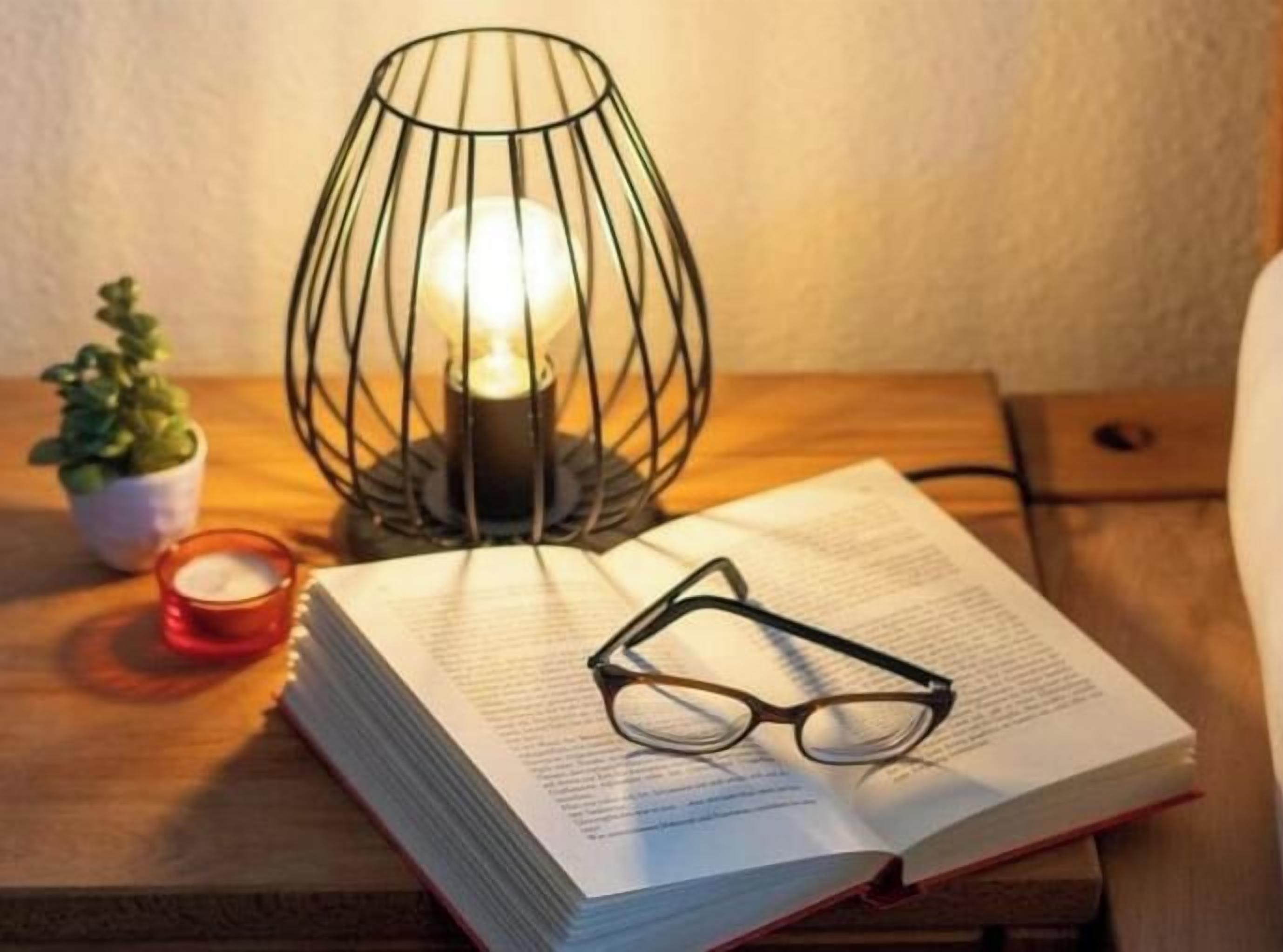 A book with glasses on top, a candle, and a wire-framed lamp on a wooden surface.