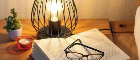 A book with glasses on top, a candle, and a wire-framed lamp on a wooden surface.