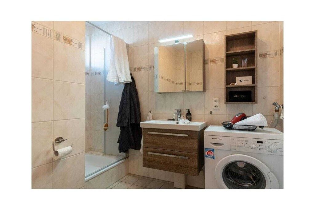 Un baño con ducha, lavamanos, espejo, toallero, estante con artículos de aseo y lavadora.