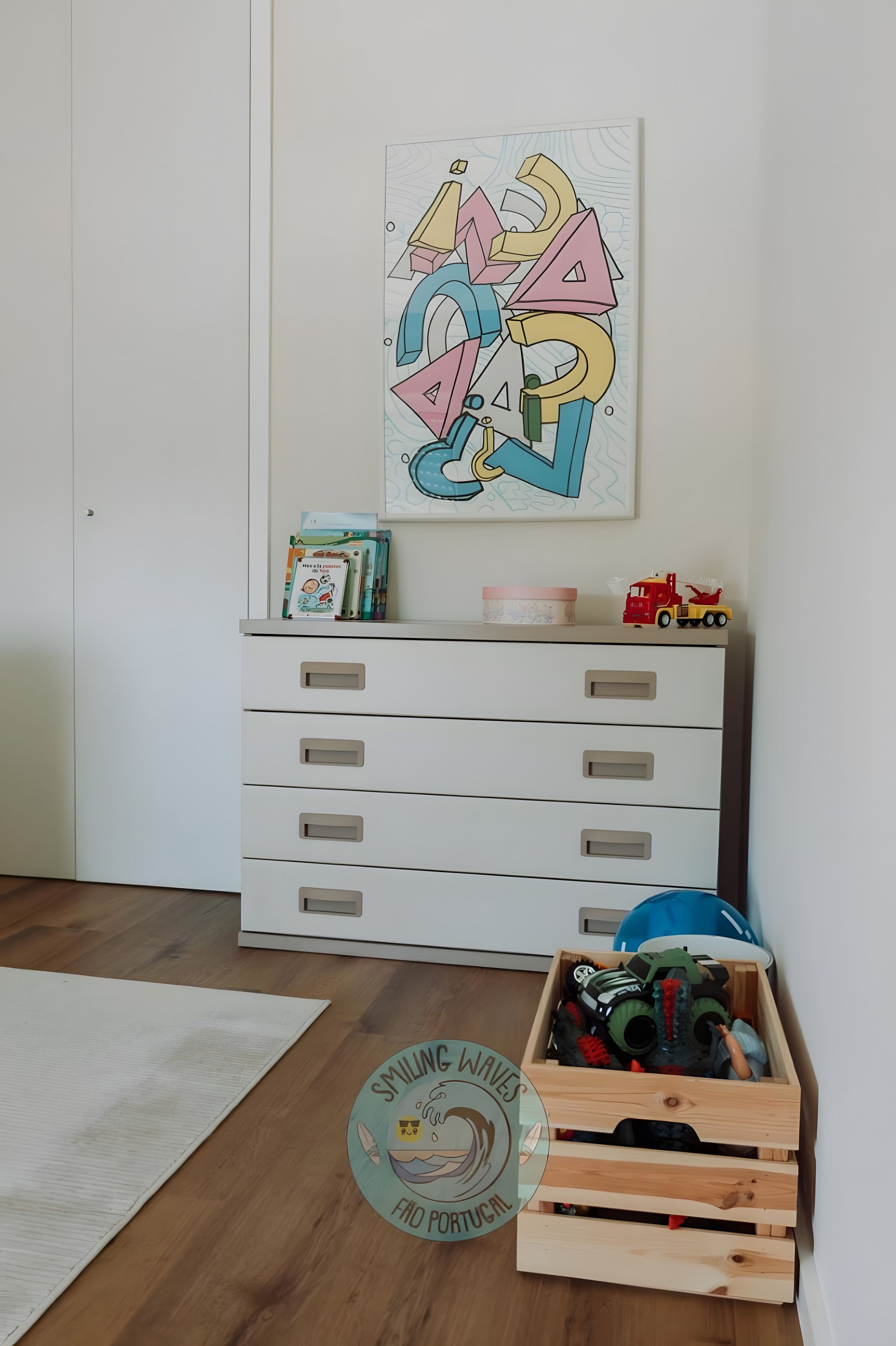 Une pièce avec une commode blanche, une caisse en bois remplie de jouets et un mur orné d’un tableau abstrait aux couleurs vives.