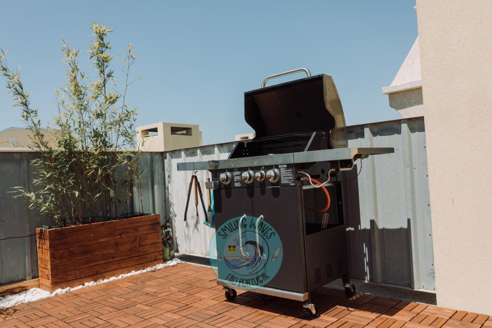 Ein tragbarer Grill auf Rädern, mit Logo und Bedienfeld, steht auf einer Ziegelterrasse mit einem hölzernen Pflanzkübel und einem Metallzaun im Hintergrund.