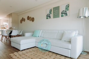 A modern living room with a white sectional sofa, a dining area with white chairs, and a kitchen in the background.