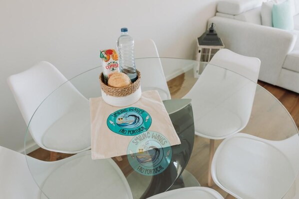 Une table ronde en verre sur laquelle repose un panier tressé contenant une bouteille et une boîte, ainsi que deux sets de table ornés d’un logo.