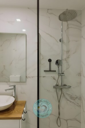 Une salle de bain avec un mur en marbre blanc, un lavabo avec un plan de travail en bois, un miroir et une douche équipée d’un pommeau de douche à main.
