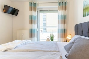 A hotel room with a bed, a flat-screen TV mounted on the wall, a window with curtains, and a view of the outside. - Holiday apartment with 2 bedrooms (Heiligenhafen)
