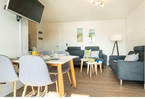 A modern living room with a dining area, a flat-screen TV mounted on the wall, and a lamp on a side table. - Holiday apartment with 2 bedrooms (Heiligenhafen)