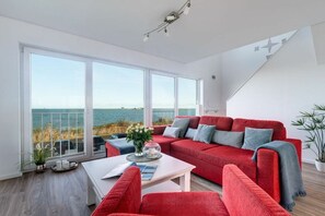 A modern living room with a red sofa, a white coffee table, and a view of the sea through large windows. - Holiday apartment with 1 bedroom (Kappeln)