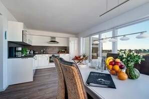 Een moderne keuken met witte kasten, een eethoek met een tafel gedekt voor twee personen, en uitzicht op het omliggende landschap door grote ramen.