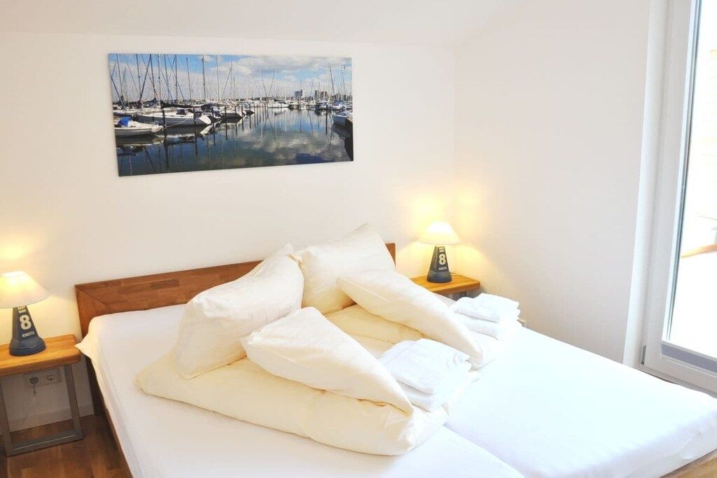 Ein Hotelzimmer mit einem Bett, zwei Nachttischlampen, einem Gemälde von einem Jachthafen und einem Fenster mit Tageslicht.