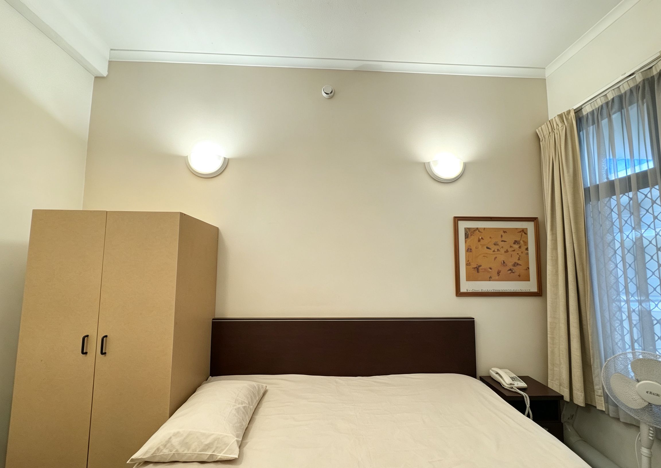 A hotel room with a bed, a wardrobe, a nightstand with a phone, and a window with curtains.
