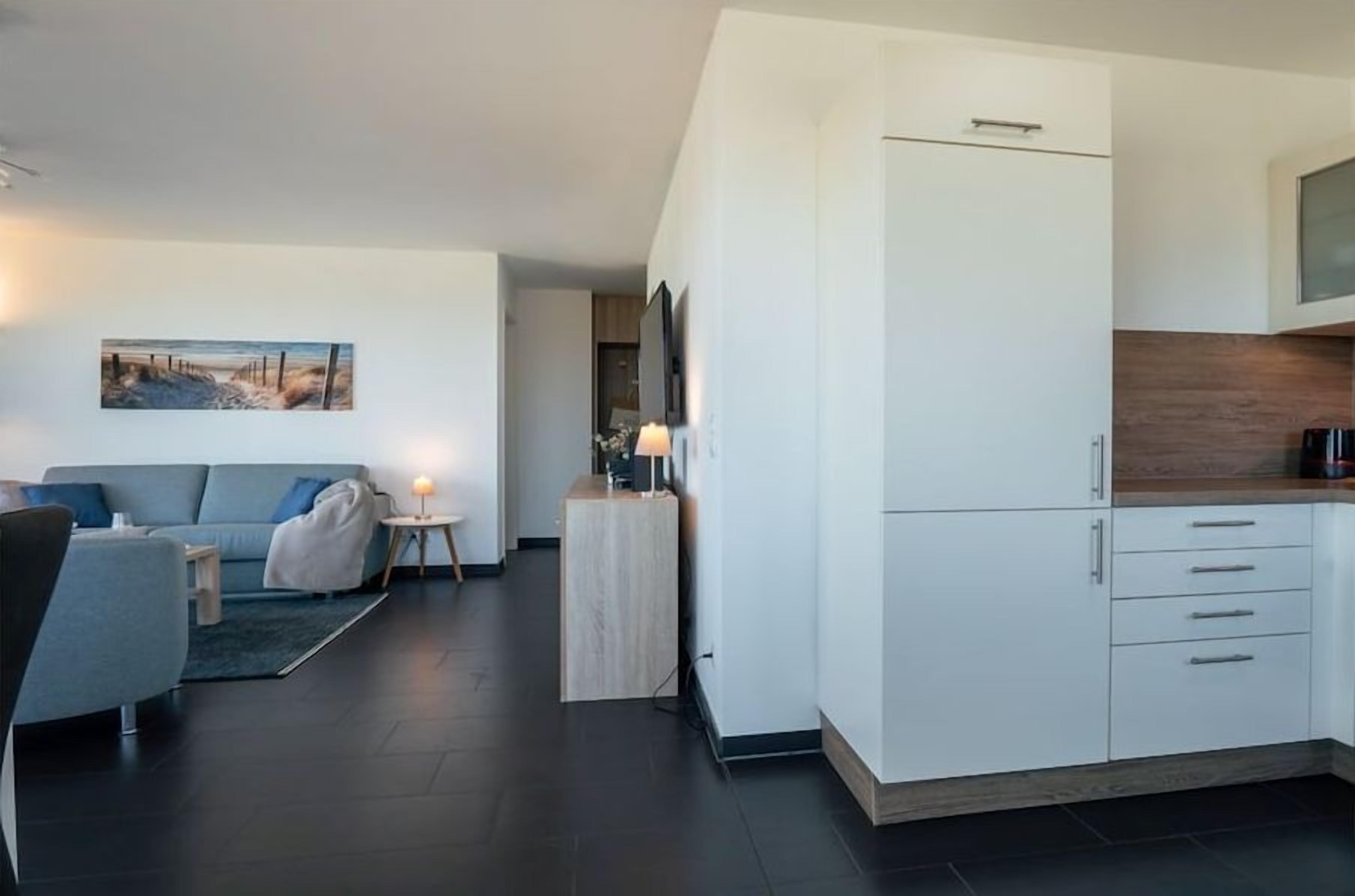 A modern living room with a sofa, a coffee table, and a small side table with a lamp. There is a kitchen area with white cabinets and a countertop.