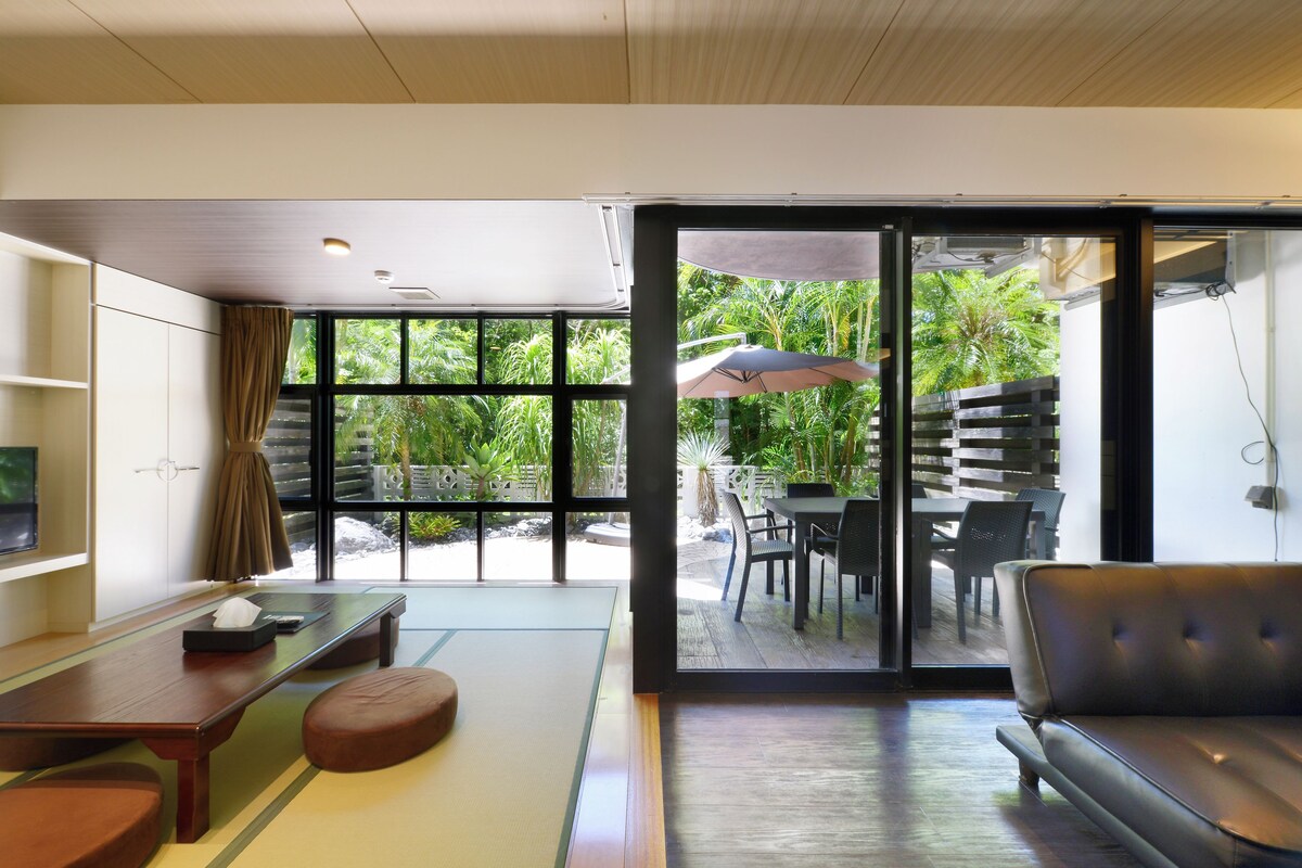 A modern living room with a sofa, a dining table with chairs, and a sliding glass door leading to an outdoor patio with a table and chairs.