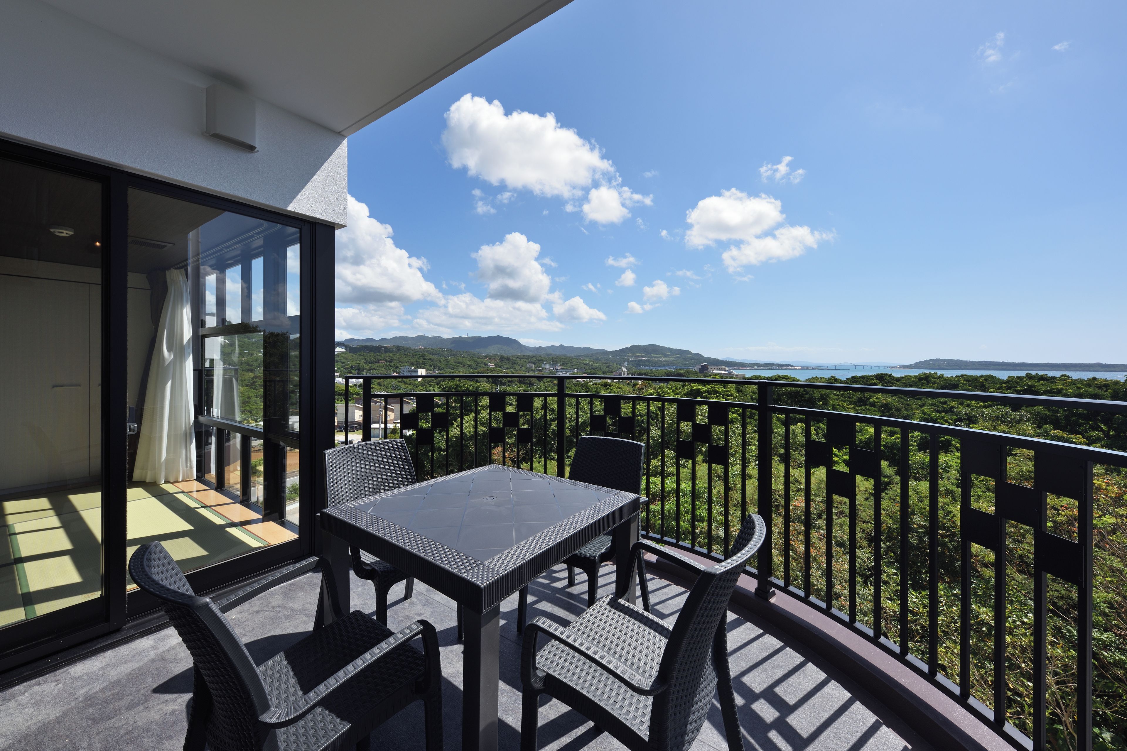 A balcony with a table and chairs, a glass door, and a view of greenery and hills.