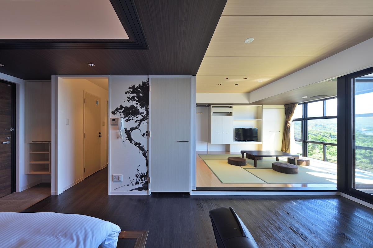 A modern hotel room with a large bed, a sitting area with tatami mats, and a view of the outdoors.