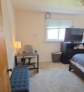 Ein Schlafzimmer mit einem Bett, einem Schreibtisch mit Stuhl, einem Fernseher, einem Fenster mit Jalousien und einer Wanduhr.