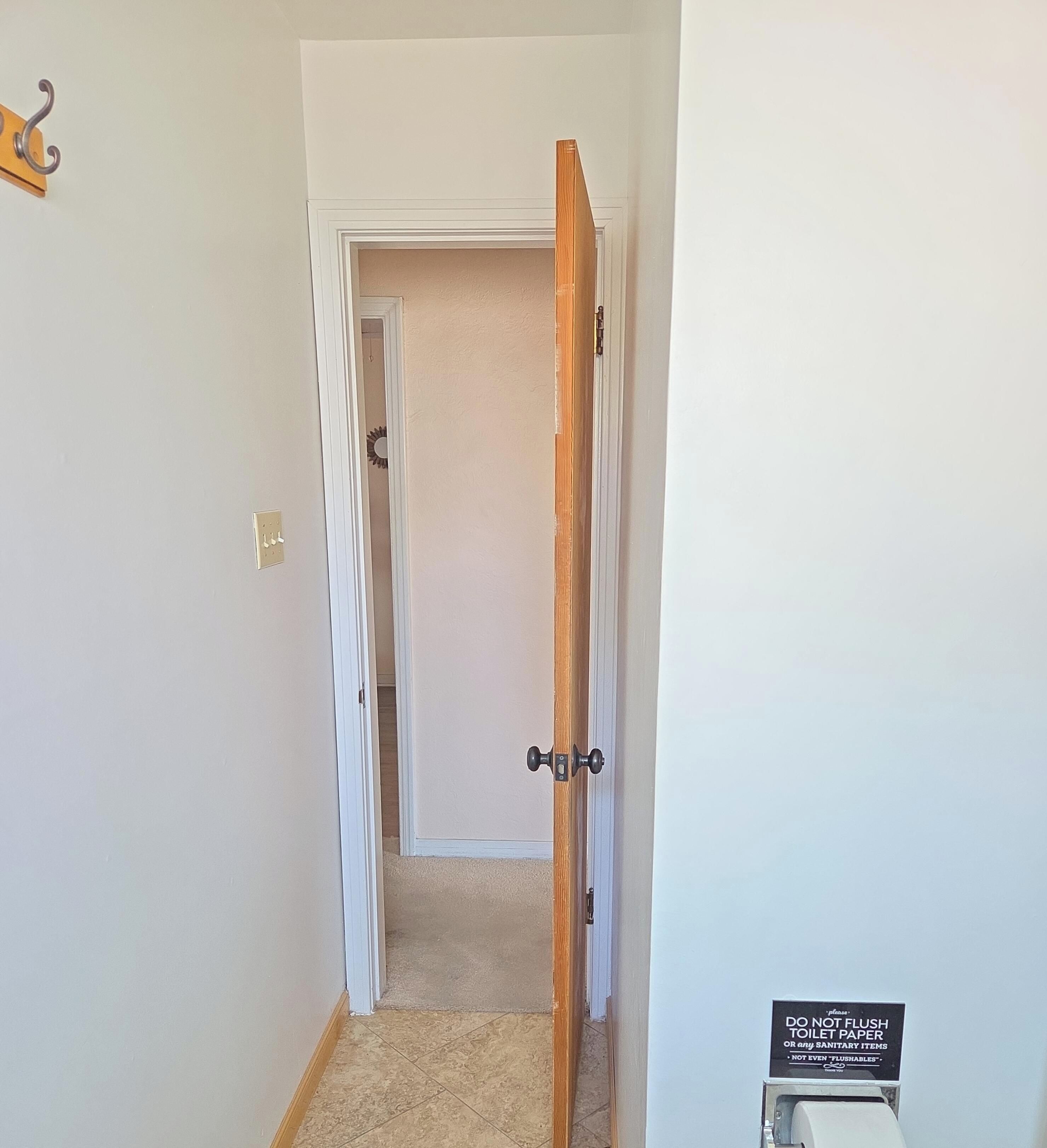 A hallway with a wooden door leading to a room, a coat hook on the wall, and a sign prohibiting flushing toilet paper.