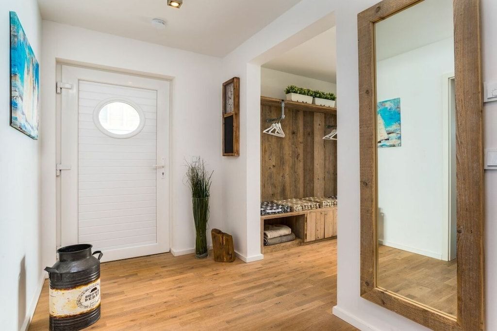A hallway with a wooden floor, a white door with a circular window, and a mirror on the right.