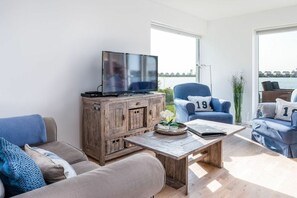 A modern living room with a television on a wooden cabinet, a coffee table with a book and a flower vase, and a sofa with blue cushions. - Cape Fred at the quayside (Kappeln)