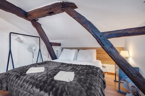 A bedroom with a sloped ceiling, exposed wooden beams, a bed with a dark blanket, white pillows, and a bedside table with a lamp. - Holiday Home "Domaine Logis Gascogne, Gîte 1": Mountain View, Pool, and Wi-Fi Included (Saint-Justin)
