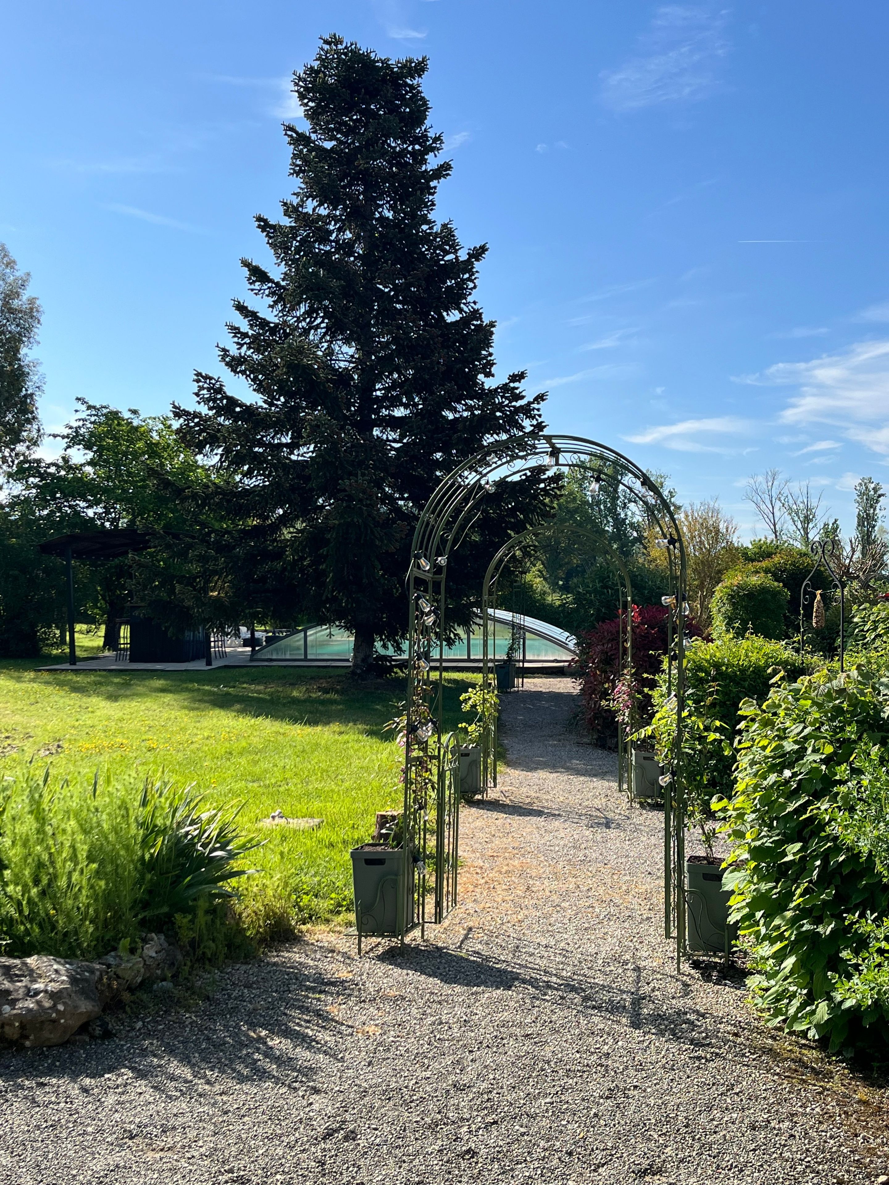 Ein Kiesweg führt zu einem Metallbogen in einem Garten mit verschiedenen Pflanzen und einem großen Baum.