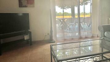 A living room with a flat-screen TV, a glass coffee table, and a sliding glass door leading to an outdoor area with a table and chairs.
