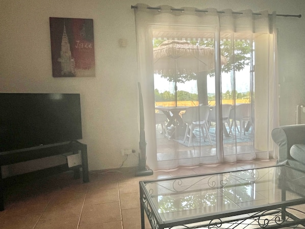 A living room with a flat-screen TV, a glass coffee table, and a sliding glass door leading to an outdoor area with a table and chairs.