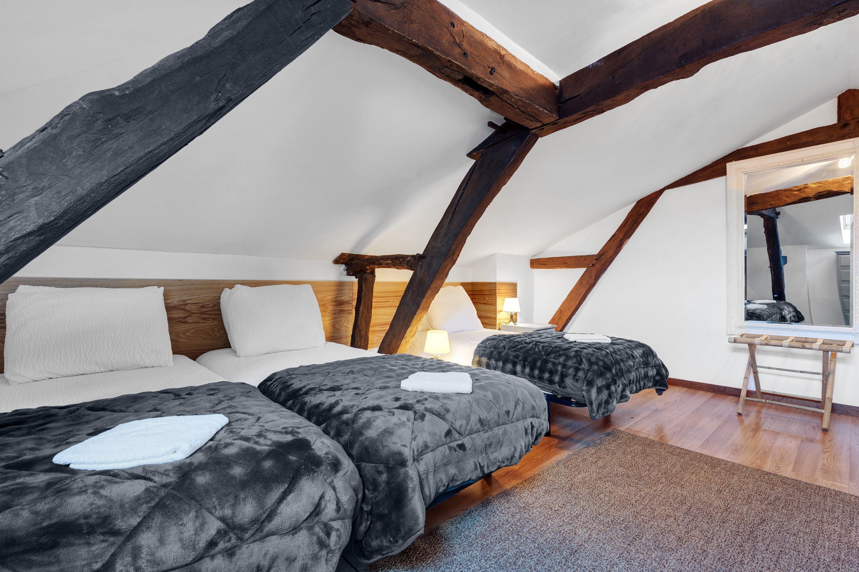 An attic bedroom with exposed wooden beams, two beds with grey bedding, a small table, and a mirror.