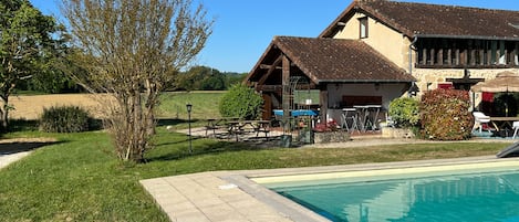 A pool area with a stone deck, a wooden building with a sloped roof, and a grassy field in the background.