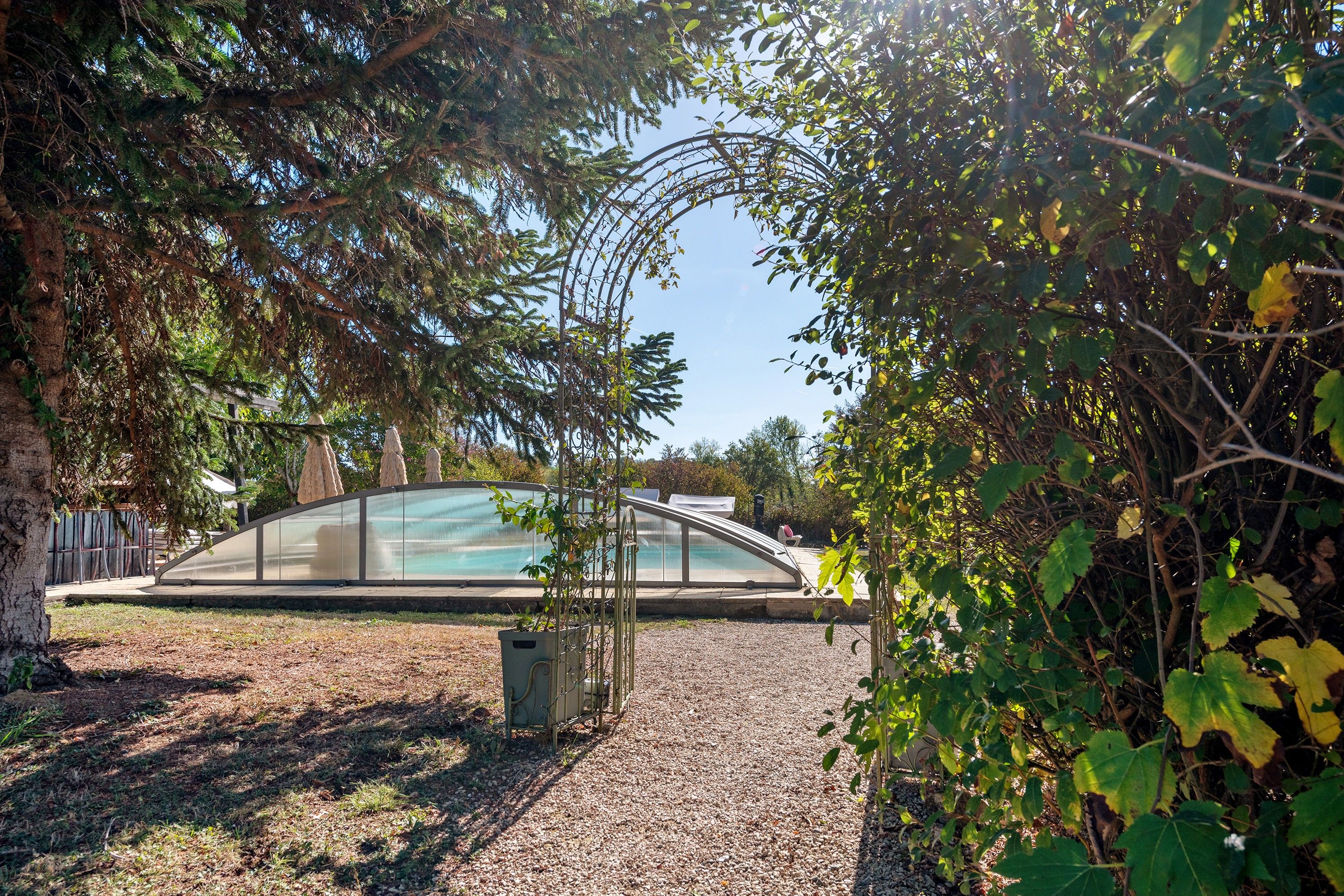 A covered outdoor area with a curved roof structure, surrounded by trees and a gravel path.