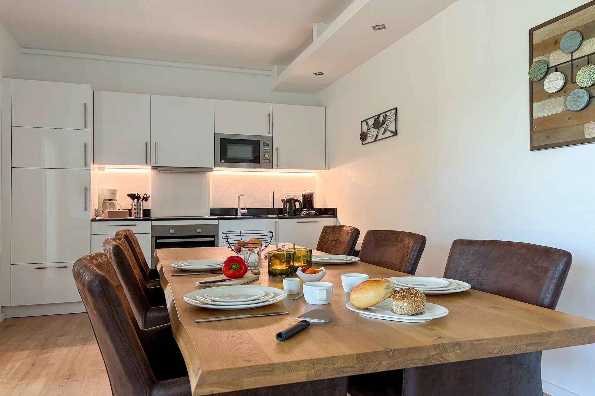 Une cuisine moderne avec des meubles blancs, un four à micro-ondes et un coin repas doté d’une table en bois mise pour deux personnes.