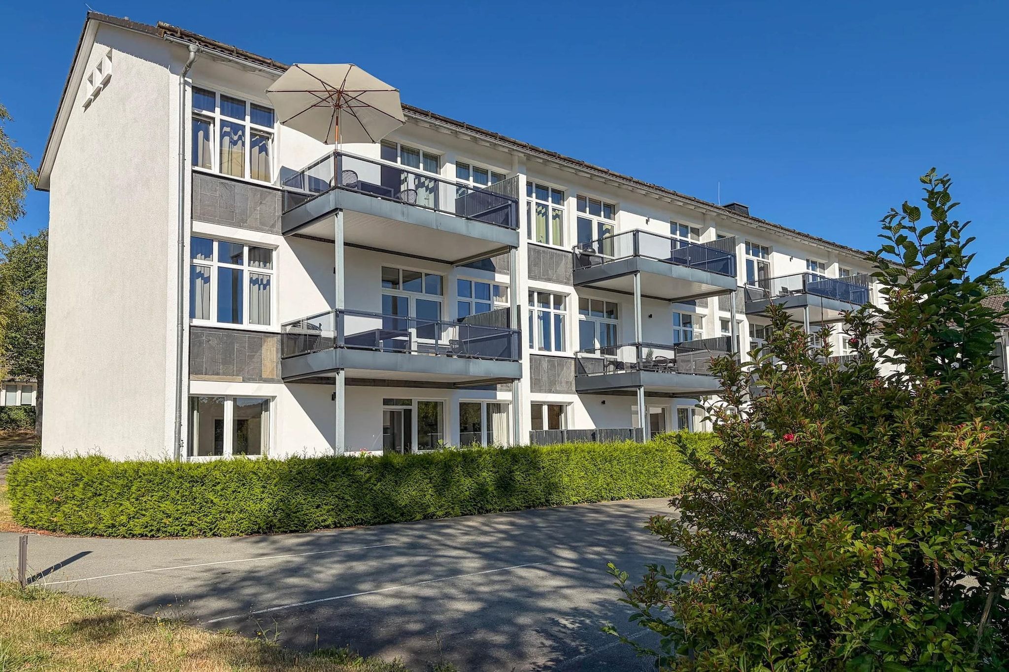 A multi-story residential building with balconies, a covered outdoor seating area, and a well-maintained hedge.