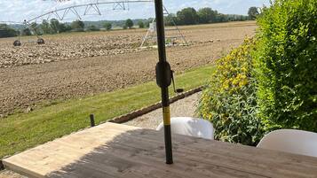 An outdoor dining area with a wooden table and white chairs, a thatched umbrella, and a view of a field.