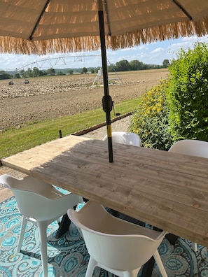 Un área de comedor al aire libre con mesa de madera y sillas blancas, un toldo de paja y vistas a un campo.