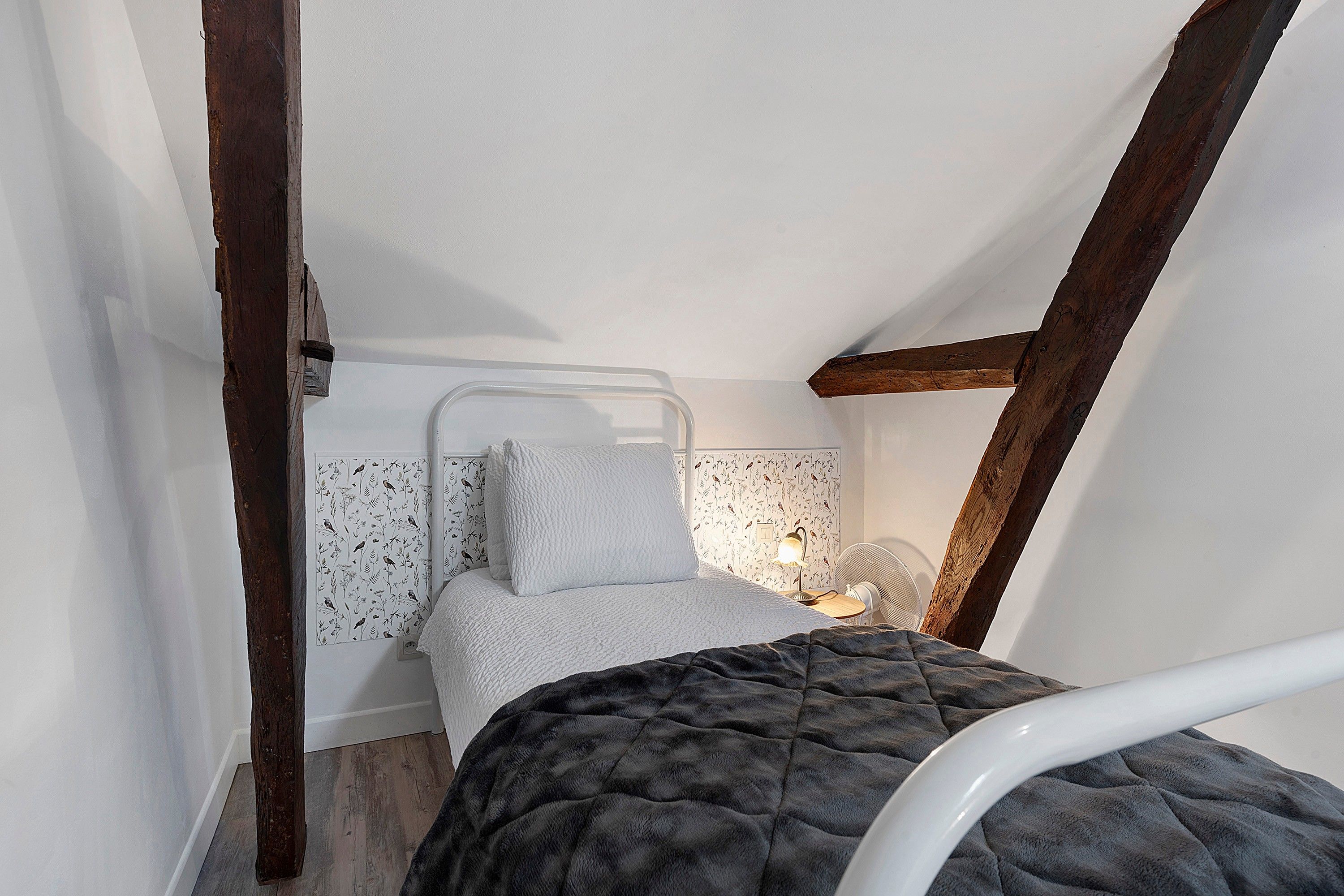 Een kleine, knusse slaapkamer met een eenpersoonsbed, een nachtkastje met lamp en een ventilator aan de muur.