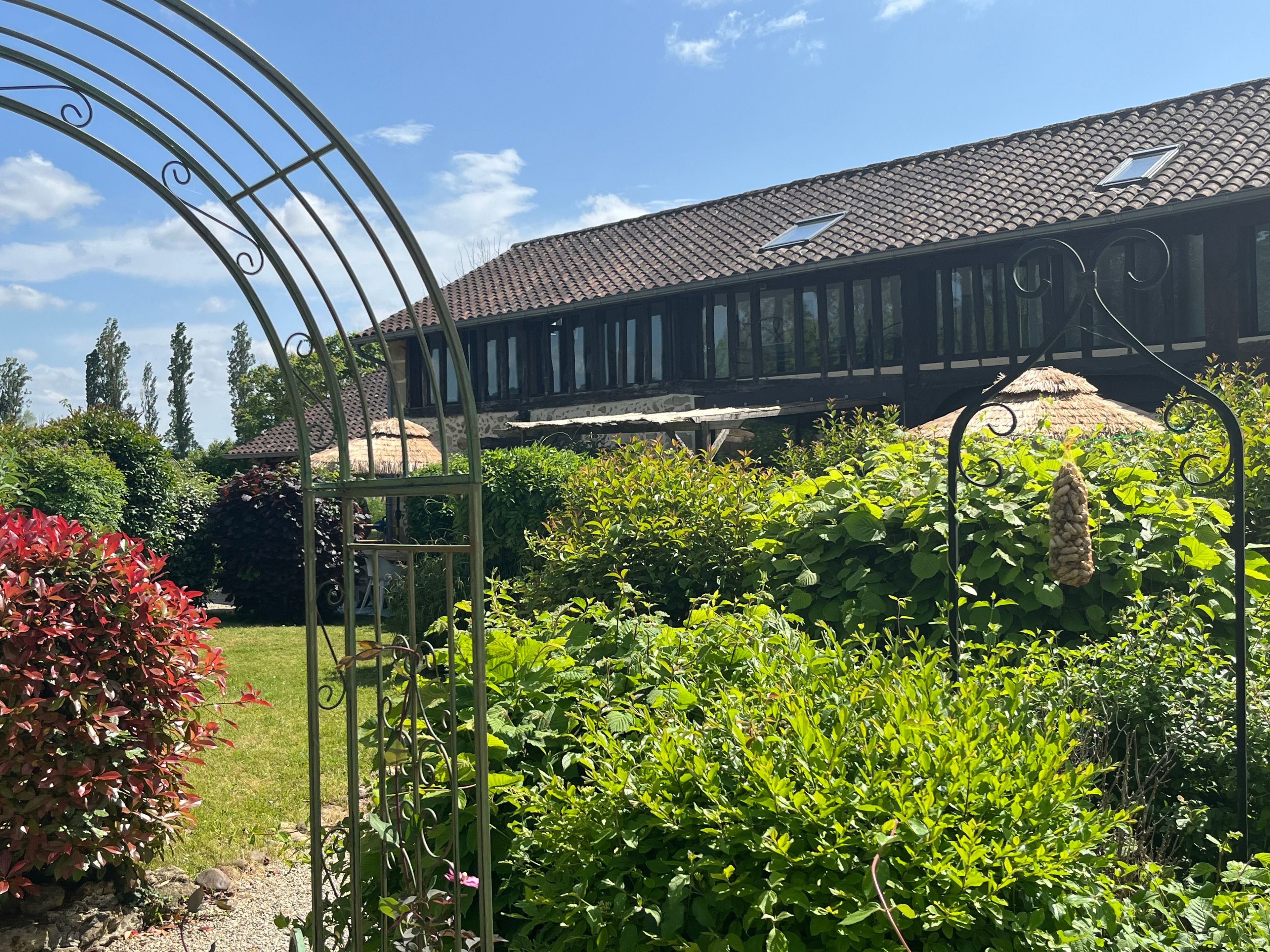 Un jardin agrémenté d’une treille en métal, une maison avec un toit en tuiles et une structure à toit de chaume.