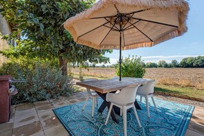 Une terrasse extérieure avec une table et des chaises, un parasol en chaume et une vue sur un champ.