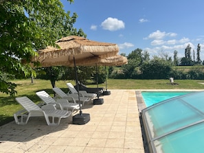 Un área junto a la piscina con sillones de descanso y sombrillas, una piscina azul cristalina y un césped.