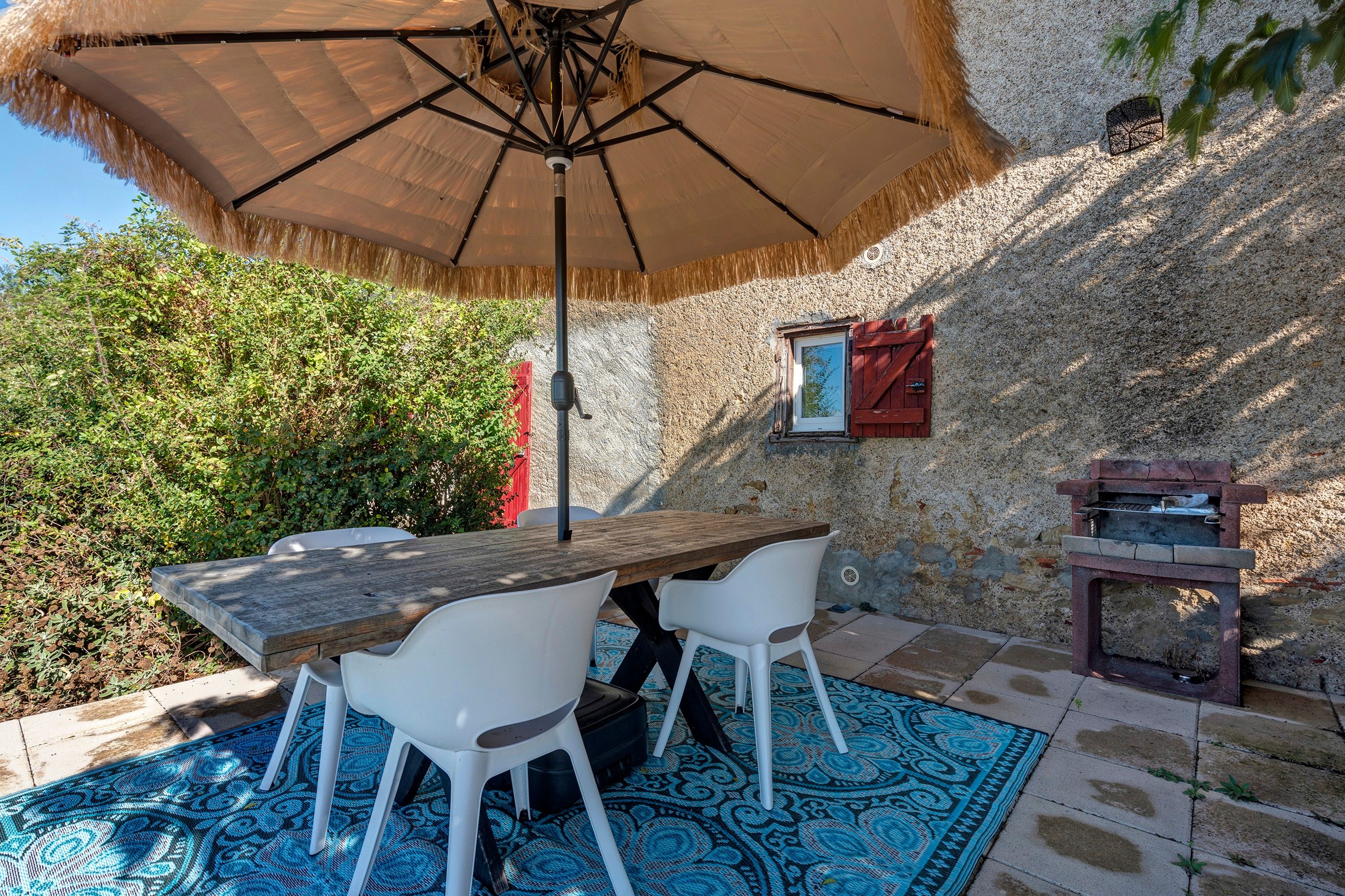 Een terras buiten met een houten tafel en witte stoelen, een rieten zonnescherm en een stenen muur.