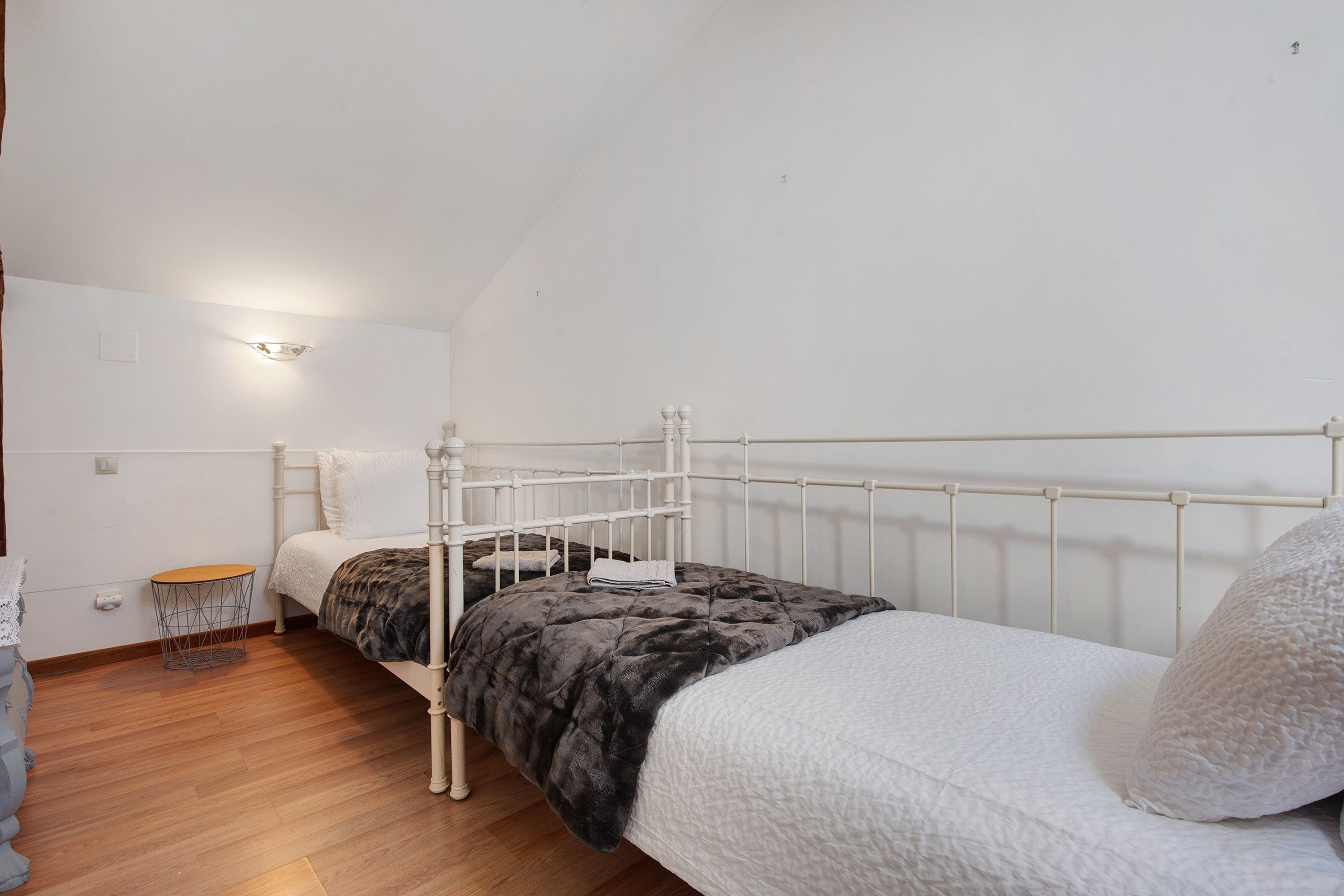 Quarto individual com cama de estrutura metálica branca, piso de madeira e uma pequena mesa redonda com uma cadeira.