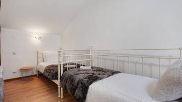 Quarto individual com cama de estrutura metálica branca, piso de madeira e uma pequena mesa redonda com uma cadeira.