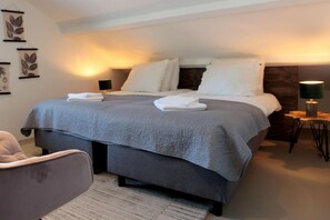 Una cama bien tendida con almohadas y una manta, una mesita de noche con una lámpara, y una silla en una habitación de hotel.