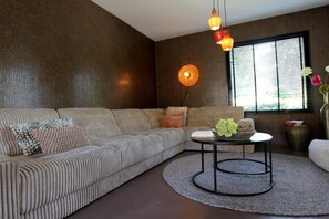 Un salon moderne avec un canapé d’angle, une table basse ronde et une grande fenêtre équipée de stores.