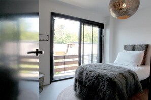 Une chambre à coucher avec un lit, une grande fenêtre avec des stores, un tapis rond et une porte dotée d’une poignée.
