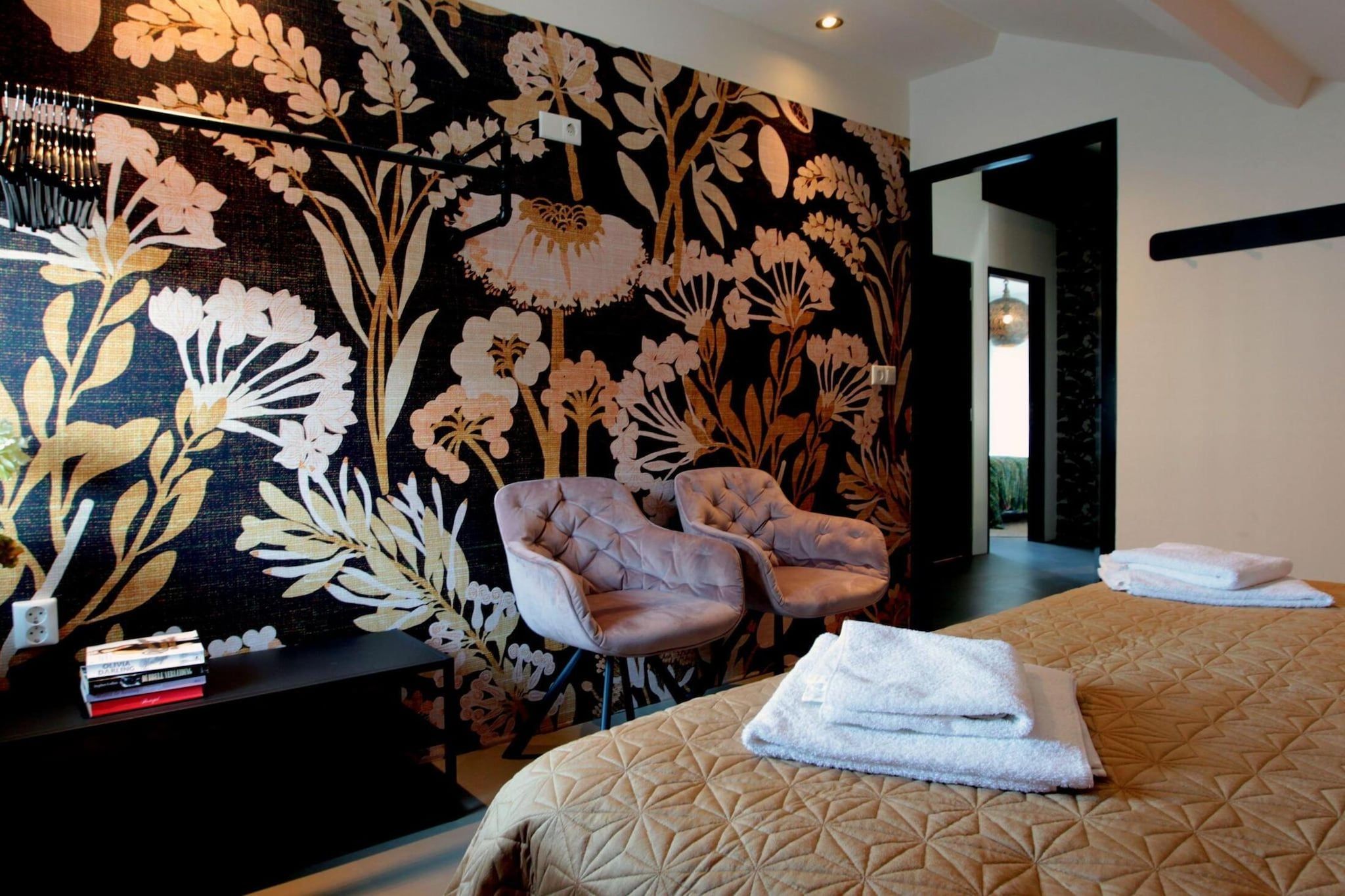 A hotel room with a large floral wall mural, a tufted armchair, a neatly made bed with a geometric patterned duvet, and a stack of folded towels.