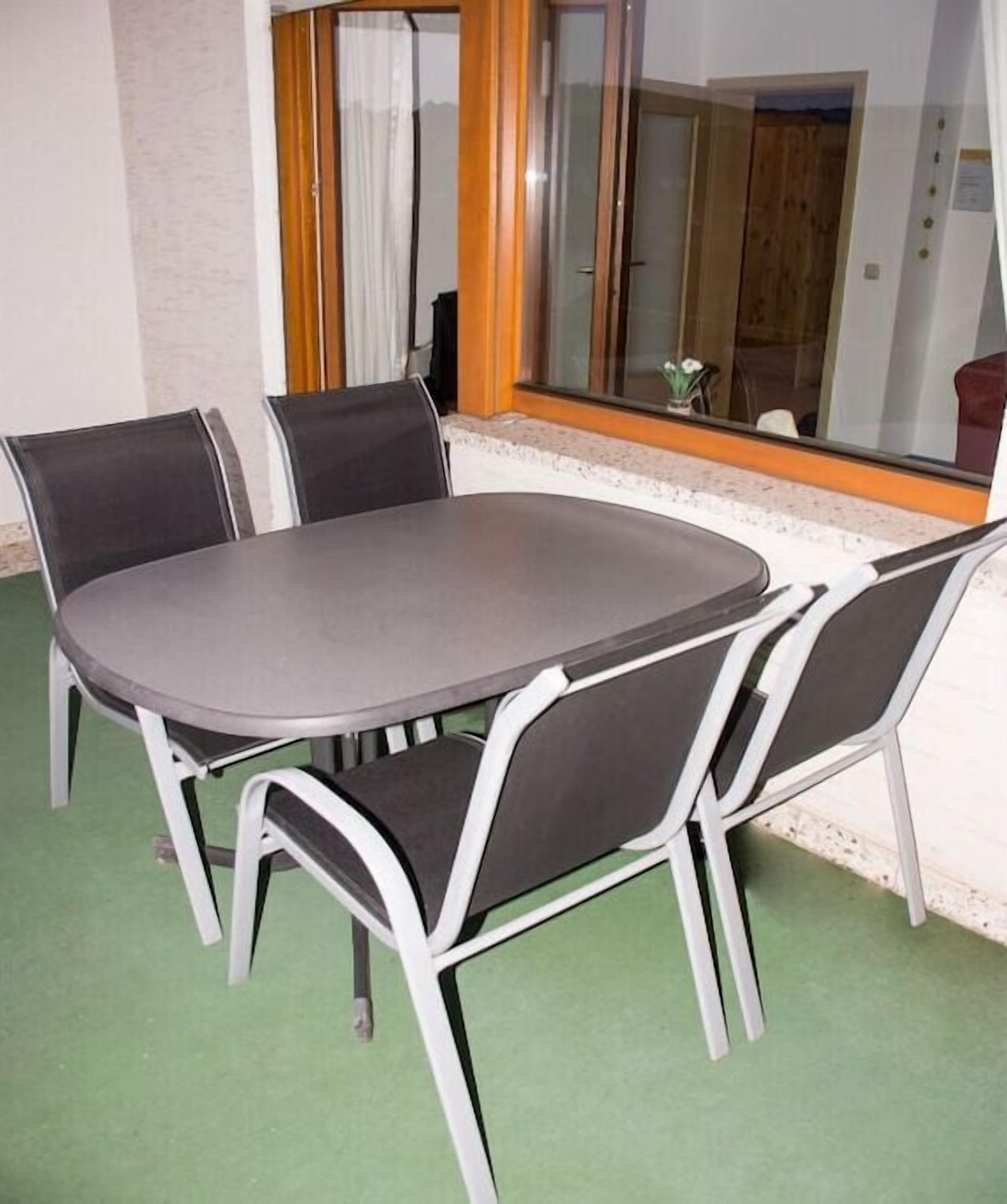 A round table with four chairs, a green carpeted floor, and a window with a view of a wooden door and a plant.