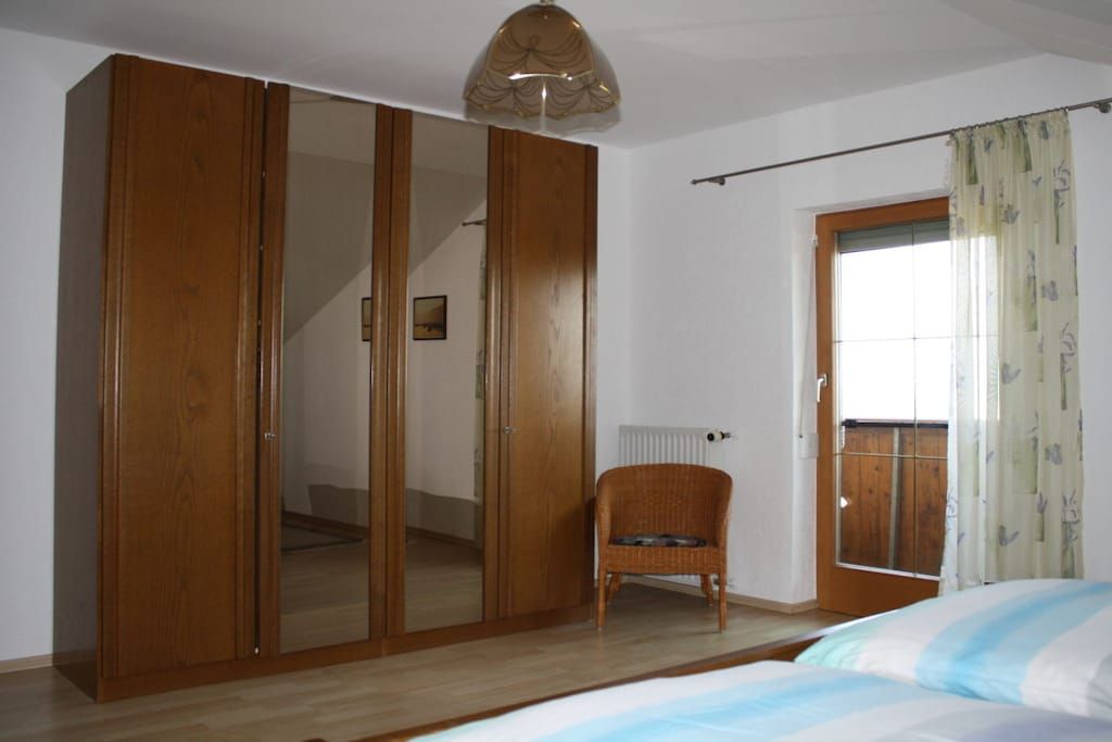 A bedroom with a wooden wardrobe, a wicker chair, a bed with striped bedding, and a window with curtains.