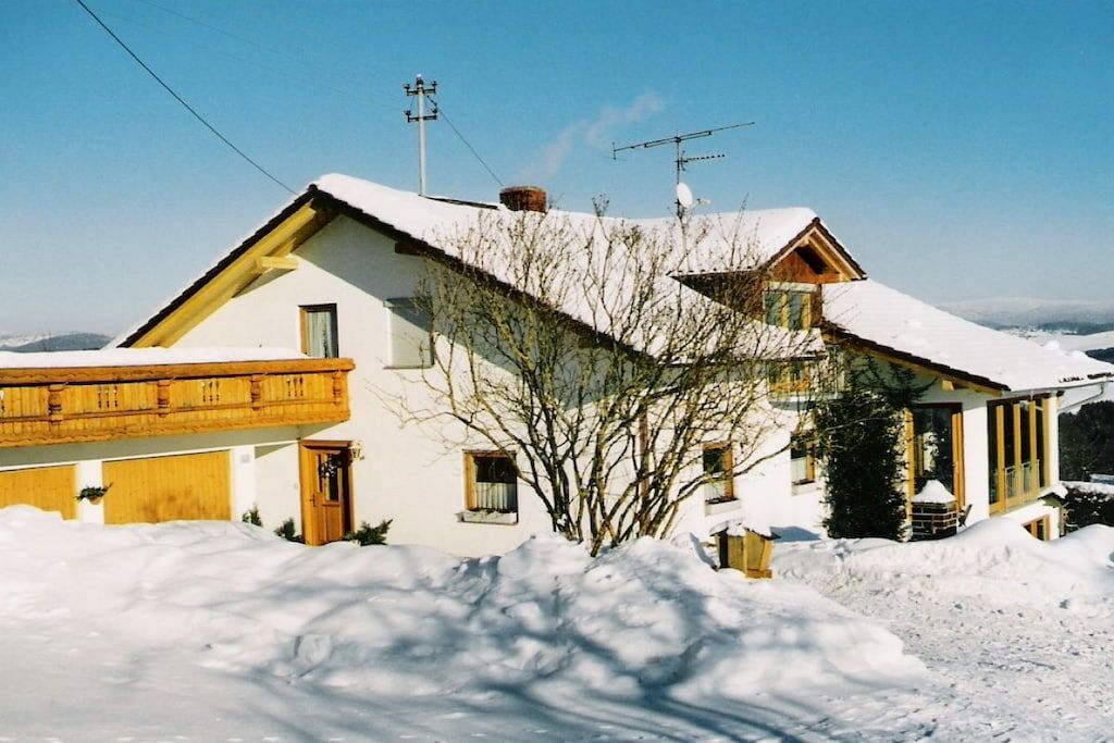 A snow-covered house with a sloped roof, a wooden deck, and a clear sky.