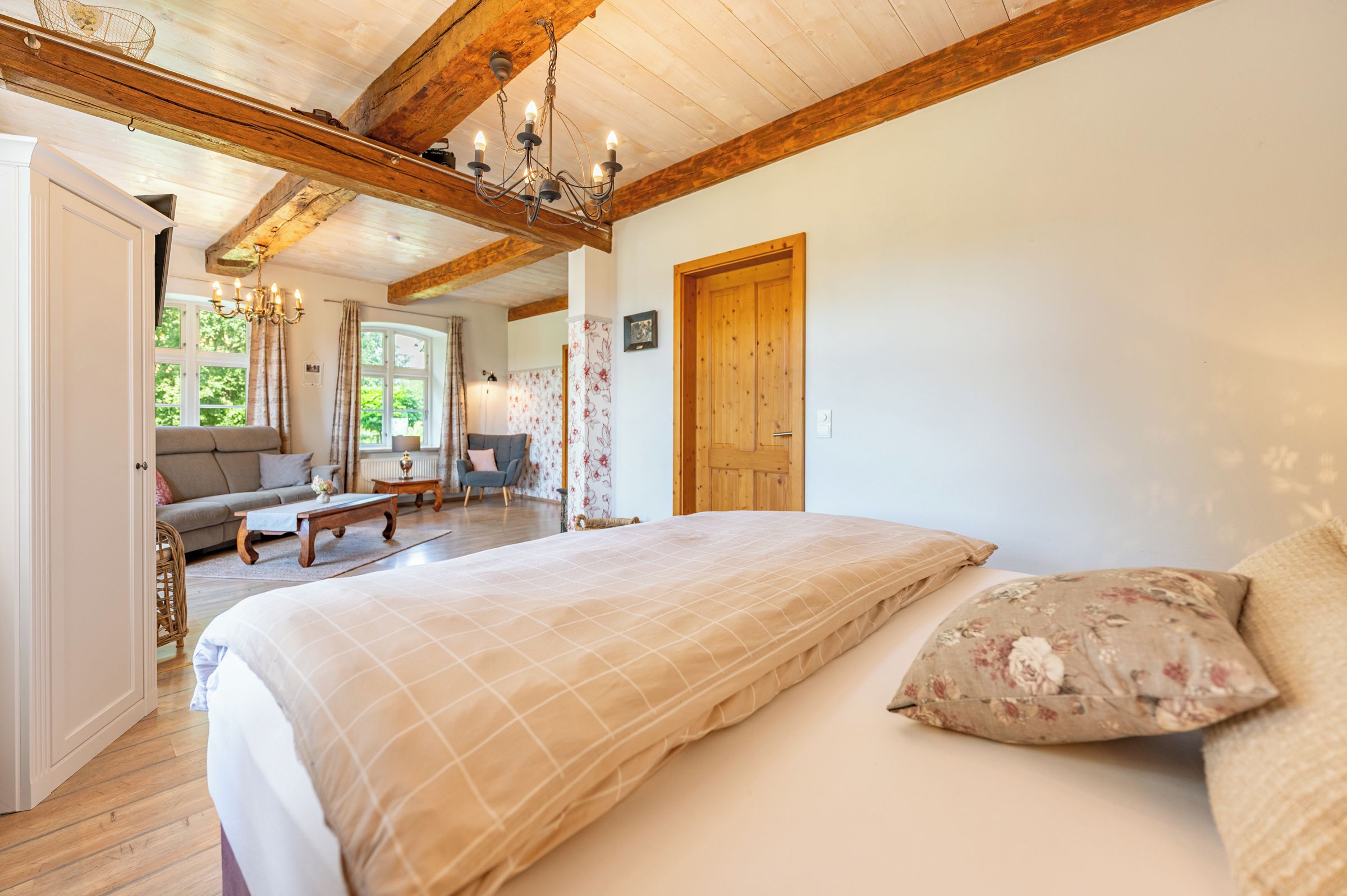 Une chambre avec un grand lit, des poutres apparentes, un lustre et une vue sur l’extérieur.