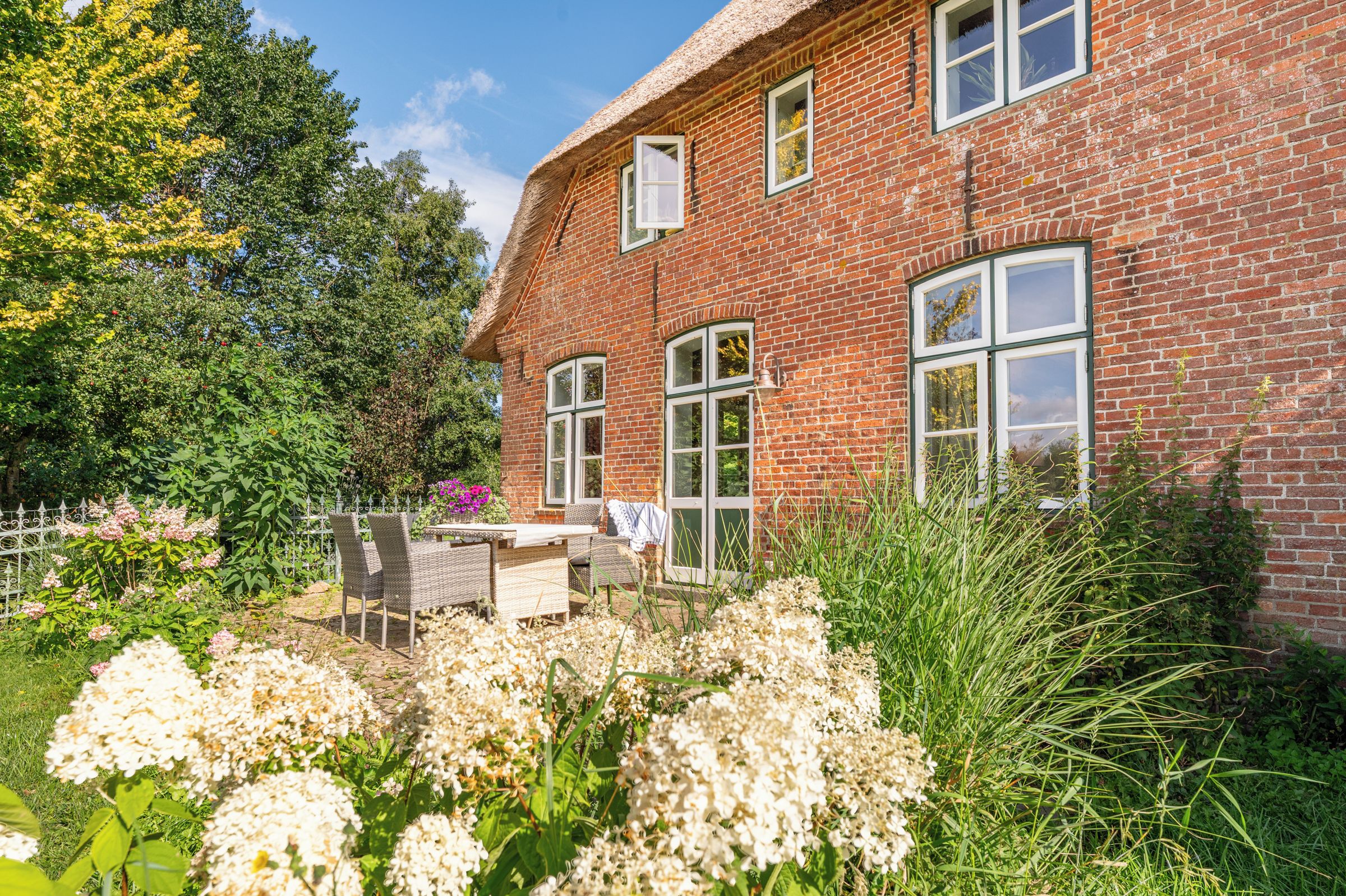A brick house with a thatched roof, a garden with white flowers, and a table with chairs outside.