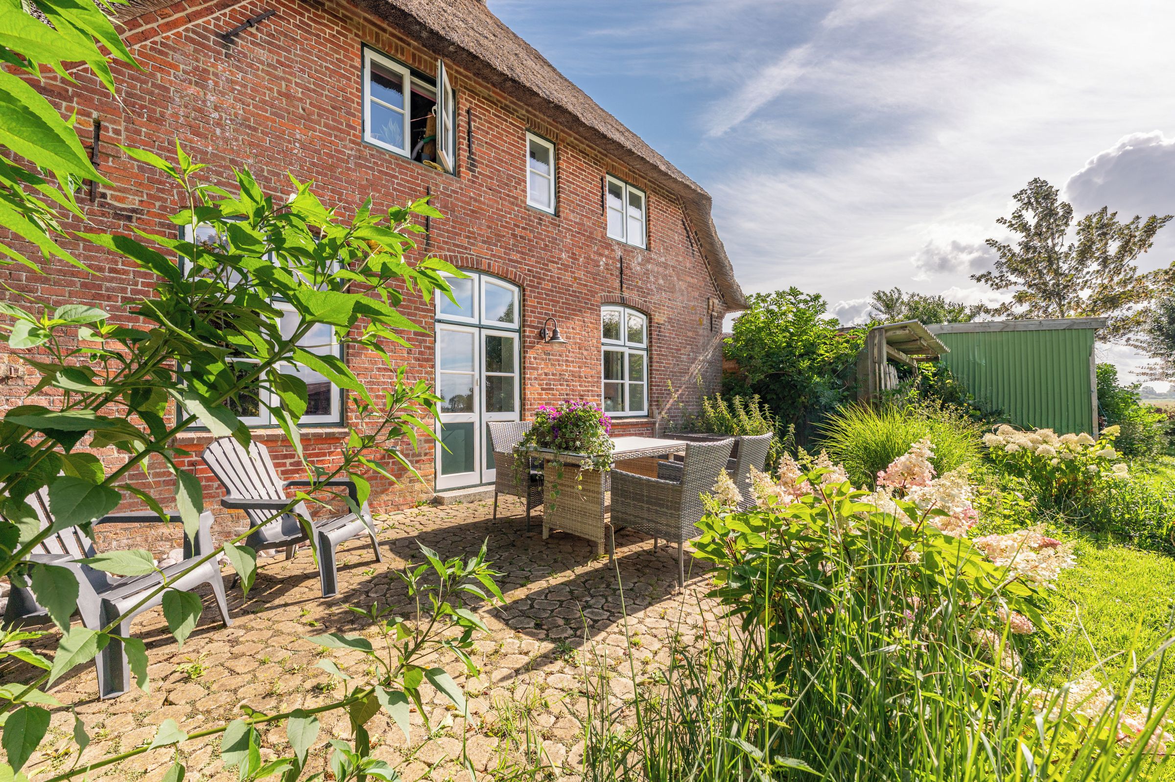 Ein Haus mit Backsteinfassade und einem Strohdach, eine gepflasterte Terrasse mit Gartenmöbeln sowie ein Garten mit verschiedenen Pflanzen und Blumen.